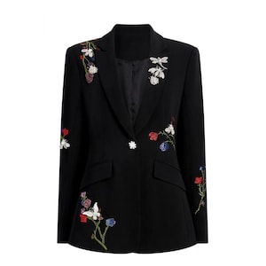 Könnte beinhalten: Ein schwarzer Blazer mit langen Ärmeln, steigendem Revers und einem Knopfverschluss. Das Kleidungsstück ist mit farbenfrohen Blumenstickereien und perlenbesetzten Insektenapplikationen, darunter eine Libelle und eine Biene, auf der Vorderseite verziert.