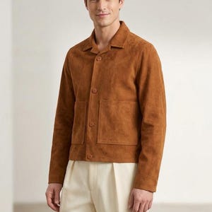 Könnte beinhalten: Braune Wildlederjacke mit Kragen und zwei Vordertaschen. Die Jacke hat einen Knopfverschluss und lange Ärmel. Das Model trägt eine cremefarbene Bundfaltenhose. Die Jacke hat einen warmen, erdigen Farbton.