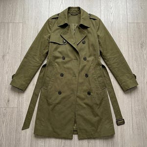 Könnte beinhalten: Ein olivgrüner zweireihiger Trenchcoat mit breitem Kragen, Schulterklappen und langen Ärmeln mit Schnallenmanschetten. Der Mantel verfügt über einen passenden Taillengürtel, zwei Reihen dunkler Knöpfe und eine Sturmklappe an der rechten Schulter.