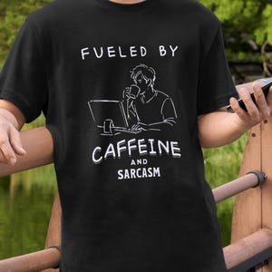 Puede incluir: Camiseta negra con el texto blanco "Fueled by Caffeine and Sarcasm". El gráfico muestra a una persona bebiendo de una taza mientras trabaja en un portátil. La camiseta es una prenda informal.