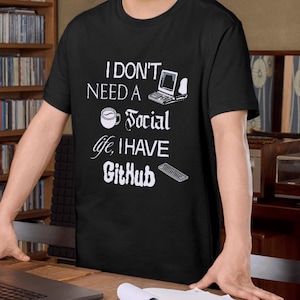 Könnte beinhalten: Schwarzes T-Shirt mit weißem Text: „I don't need a social life, I have GitHub.“ Das Design zeigt eine Kaffeetasse, einen Computer und eine Tastatur. Die Person steht vor einem Bücherregal und einem Schreibtisch.