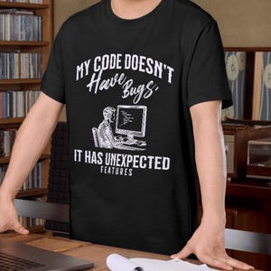 May include: Black t-shirt with the text "MY CODE DOESN'T Have Bugs, IT HAS UNEXPECTED FEATURES" in white. The design includes a graphic of a person working on a computer. The shirt is worn by a person.