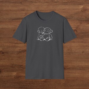May include: Dark grey t-shirt with a white outline illustration of a child embracing a monkey. The design is positioned centrally on the chest. The shirt has short sleeves and a crew neck.