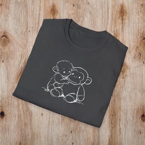 May include: Dark grey t-shirt with a white outline illustration of two monkeys hugging. The design is centrally placed on the folded shirt, which rests on a wooden surface. The t-shirt has a crew neck.