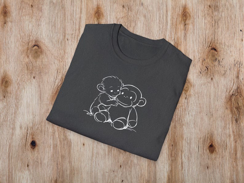 May include: Dark grey t-shirt with a white outline illustration of two monkeys hugging. The design is centrally placed on the folded shirt, which rests on a wooden surface. The t-shirt has a crew neck.