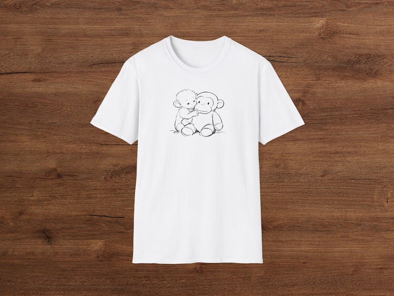 May include: White t-shirt with a black and white illustration of a child hugging a monkey. The design is centered on the front. The shirt has a crew neck and short sleeves. The background is a dark wood grain.