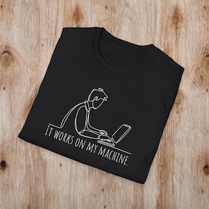 以下が含まれることがあります： ノートパソコンで作業している人の白い線画が描かれた黒いTシャツ。「IT WORKS ON MY MACHINE」の文字が下に白でプリントされています。木製の表面にシャツが表示されています。