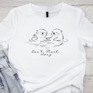 May include: A white t-shirt featuring a black line art graphic of two otters holding hands while floating in water. One otter holds a small flower. Below the illustration, the text reads "Don't Float Away" in a black cursive script font.