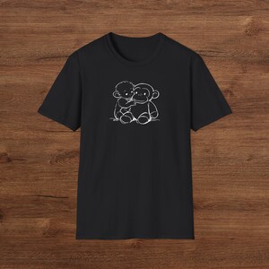 May include: Black t-shirt with a white illustration of a child embracing a monkey. The design is a simple, cartoon-style drawing of the two figures. The t-shirt has a crew neck and is made of a soft fabric.