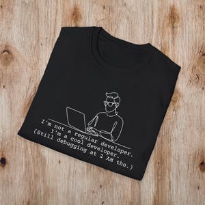 Op de afbeelding: Zwart T-shirt met een witte lijntekening van een persoon die op een laptop werkt. De tekst op het shirt luidt: "Ik ben geen gewone ontwikkelaar. Ik ben een coole ontwikkelaar. (Nog steeds aan het debuggen om 2 uur 's ochtends.)"