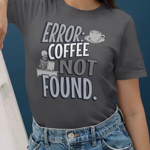 Può includere: T-shirt grigio scuro con la scritta "ERROR: COFFEE NOT FOUND." Il design include una tazza di caffè, uno scheletro a una scrivania e il testo in bianco e grigio. Un capo d'abbigliamento casual.