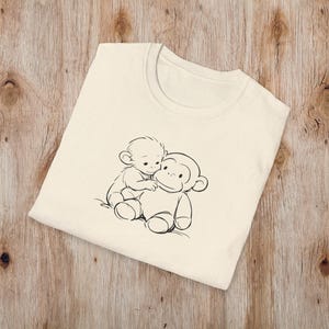 May include: Cream-coloured t-shirt with a black and white illustration of two cartoon monkeys. One monkey is sitting on top of the other. The t-shirt is made of a soft material and has a crew neck.