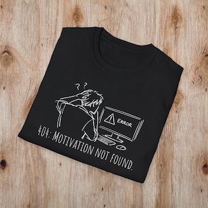 May include: Black t-shirt featuring a white graphic of a person looking at a computer screen with an error message. The text on the shirt reads "404: MOTIVATION NOT FOUND."