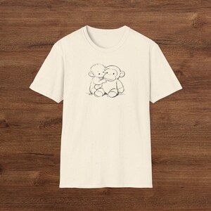 May include: Cream-coloured t-shirt with a black and white illustration of a small figure hugging a monkey. The graphic is centred on the front. The shirt is made of a soft material.