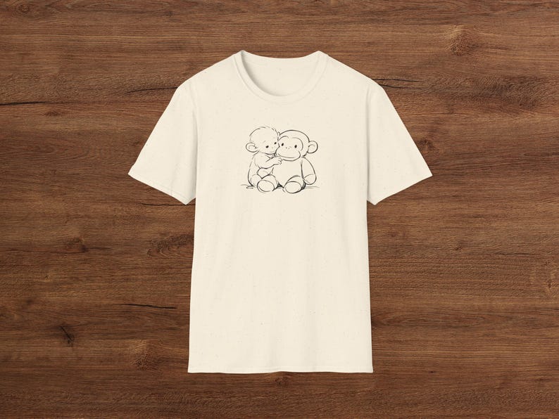 May include: Cream-coloured t-shirt with a black and white illustration of a small figure hugging a monkey. The graphic is centred on the front. The shirt is made of a soft material.