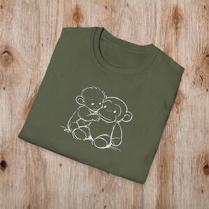 May include: An olive green t-shirt with a white outline illustration of two cartoon monkeys hugging. The t-shirt is folded on a wooden surface. The design is simple and playful, suitable for casual wear.