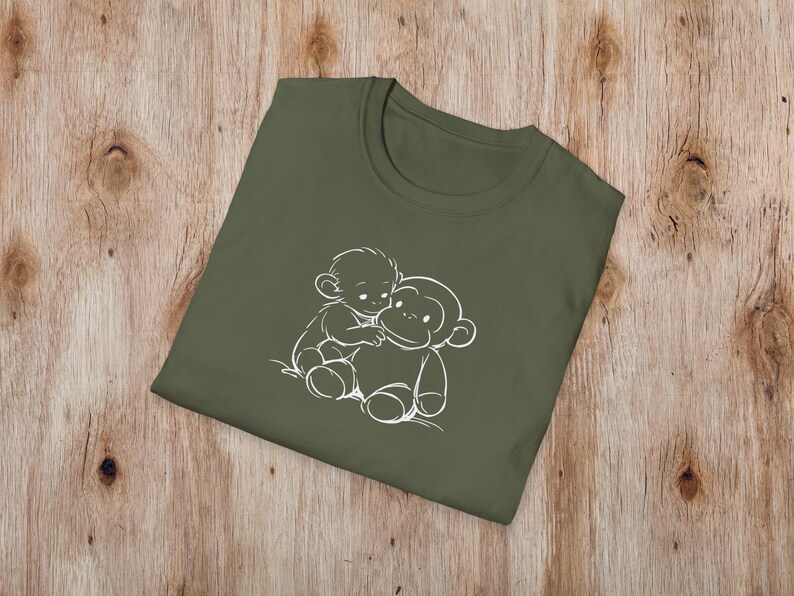May include: An olive green t-shirt with a white outline illustration of two cartoon monkeys hugging. The t-shirt is folded on a wooden surface. The design is simple and playful, suitable for casual wear.