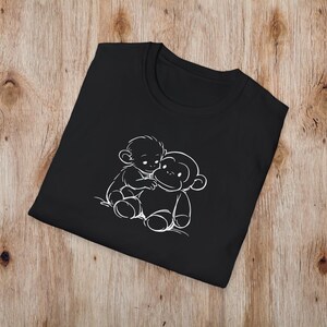May include: Black t-shirt with a white outline illustration of two cartoon monkeys hugging. The design is centrally placed on the folded shirt, which is on a wooden surface. The t-shirt has a crew neck.