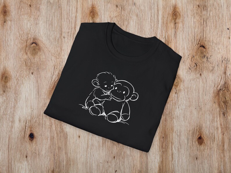 May include: Black t-shirt with a white outline illustration of two cartoon monkeys hugging. The design is centrally placed on the folded shirt, which is on a wooden surface. The t-shirt has a crew neck.