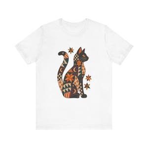 Może przedstawiać: Biały t-shirt z krótkim rękawem z grafiką siedzącego kota ozdobionego patchworkowym wzorem gwiazd, księżyców, dyń i geometrycznych kształtów w odcieniach pomarańczu, czerni i kremu, w towarzystwie trzech małych ikon w kształcie gwiazdek.