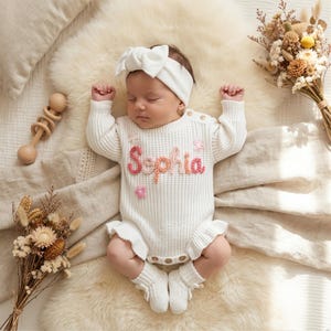 May include: A newborn baby wearing a white knit romper with the name "Sophia" embroidered in pink and peach. The outfit includes matching socks and a headband. The baby is lying on a fluffy white rug, with dried flowers and a wooden rattle nearby.