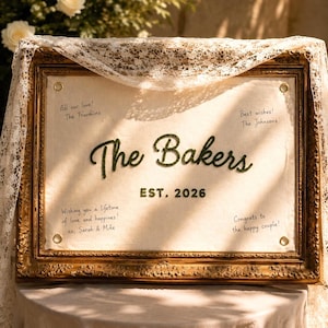 May include: A rectangular off-white fabric sign with dark green embroidered text reading The Bakers EST. 2026, surrounded by handwritten messages in a gold ornate frame draped with white lace. Four metal grommets secure the fabric to the frame.
