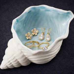 May include: A white ceramic seashell-shaped dish with a light blue interior holds gold jewelry. The jewelry includes two flower-shaped stud earrings, two teardrop earrings, and two gold rings with clear stones.