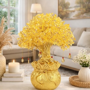 May include: A golden decorative tree sculpture with a money bag base. The tree's foliage is made of yellow crystals, and it has hanging ornaments. The sculpture is on a white surface, with a candle and vase nearby. The overall theme is wealth and prosperity.