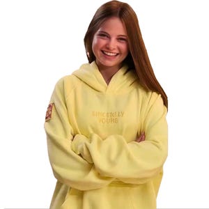 May include: A pale yellow hoodie with the embroidered text "Sincerely Yours" across the chest. The hoodie has a hood and a small patch on the left sleeve. The person is smiling.