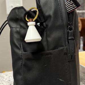 May include: Black nylon shoulder bag with a small white and brown charm hanging from a gold ring. The bag has a front pocket and a zipper closure. A small tag with the letters "ROG" is visible.