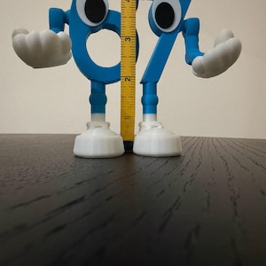 May include: A blue number 67 figurine with cartoon eyes, arms, and hands. The figurine is approximately 13 cm tall, with white feet and a yellow ruler in the background. The figurine is standing on a dark brown surface.