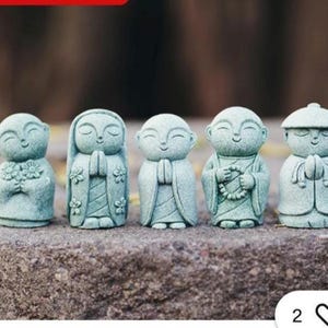May include: Five small, pale green statues of smiling figures in different poses. The statues are on a stone surface, with a blurred background. Each figure has a unique design, including flowers, prayer hands, and a hat.