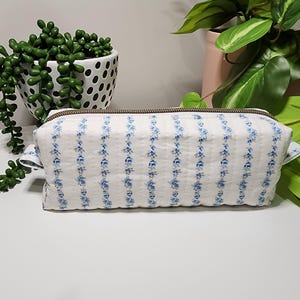 May include: A rectangular quilted fabric pouch featuring a white background with vertical stripes of small blue floral patterns. The pouch has a brown zip closure along the top edge and small fabric tabs on both sides of the rectangular structure.