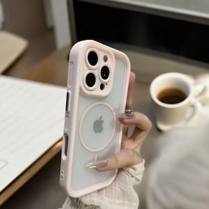 Può includere: Una custodia trasparente per telefono con bordo rosa chiaro e protezione dell'obiettivo della fotocamera. La custodia presenta un design circolare con il logo Apple e un anello magnetico. La custodia è progettata per uno smartphone.