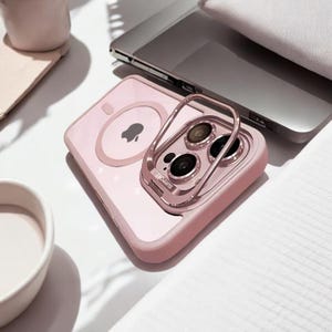 Op de afbeelding: Een roze telefoonhoesje met een doorzichtige achterkant en een metalen ringhouder. De hoes heeft een ingebouwde magnetische oplaadfunctie en een verhoogde rand rond de camera. Het Apple-logo is zichtbaar op de achterkant.