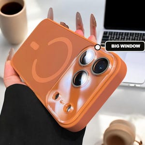 Könnte beinhalten: Orangefarbene Handyhülle mit einer transparenten Kameraobjektivabdeckung. Die Hülle hat ein Smiley-Gesicht-Design auf der Rückseite. Die Worte "BIG WINDOW" sind im Hintergrund sichtbar.