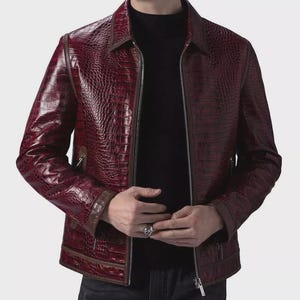 Peut inclure: Veste en cuir bordeaux avec une texture peau de crocodile. La veste a une fermeture éclair, un col et des bordures marron sur les poignets et les poches. La veste est portée sur un col roulé noir.