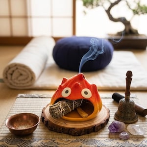 May include: A colourful ceramic incense holder in orange and yellow, shaped like a cartoon character, with a bundle of sage inside. A small copper bowl, bell, and crystals are nearby, suggesting a spiritual theme. A bonsai tree and a blue meditation cushion are in the background.