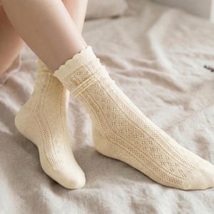 May include: Cream-colored, open-knit socks with a scalloped edge. The socks have a delicate, patterned design and are worn on a bed. The socks are a light, neutral color, and the overall aesthetic is soft and comfortable.