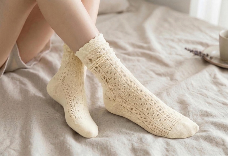 May include: Cream-colored, open-knit socks with a scalloped edge. The socks have a delicate, patterned design and are worn on a bed. The socks are a light, neutral color, and the overall aesthetic is soft and comfortable.