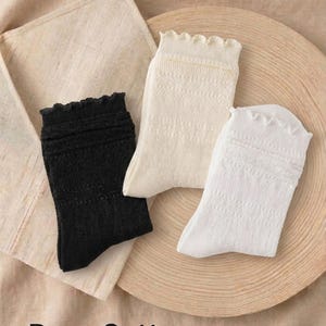 May include: Three pairs of socks in black, cream, and white, each featuring a ruffled cuff and a subtle knit texture. The text on the image reads: Pure Cotton, Delicate & Skin-friendly / Natural Antibacterial. 100% COTTON, with two circular icons.