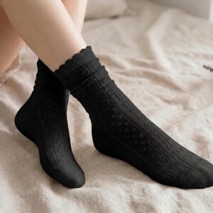 May include: A pair of black socks with a scalloped edge and a textured, openwork design. The socks are knee-high and made of a soft material. The socks are displayed on a bed with a neutral-colored sheet.