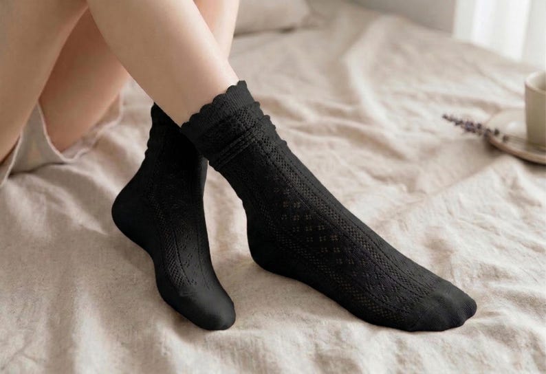 May include: A pair of black socks with a scalloped edge and a textured, openwork design. The socks are knee-high and made of a soft material. The socks are displayed on a bed with a neutral-colored sheet.