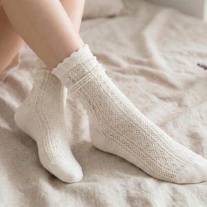 May include: A pair of off-white, open-knit socks with a scalloped trim. The socks feature a delicate diamond pattern and are worn on a bed with a neutral-colored sheet. The socks are knee-high and have a textured appearance.
