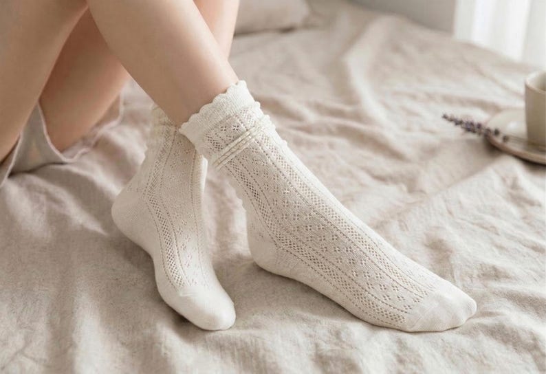 May include: A pair of off-white, open-knit socks with a scalloped trim. The socks feature a delicate diamond pattern and are worn on a bed with a neutral-colored sheet. The socks are knee-high and have a textured appearance.
