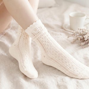 May include: A pair of cream-colored, open-knit socks with a scalloped trim. The socks feature a diamond pattern and are knee-high. A white mug and a bundle of dried lavender are in the background.