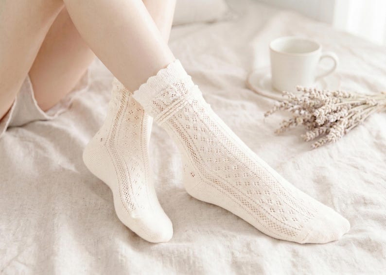 May include: A pair of cream-colored, open-knit socks with a scalloped trim. The socks feature a diamond pattern and are knee-high. A white mug and a bundle of dried lavender are in the background.