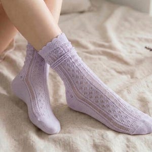 May include: A pair of lavender socks with a delicate openwork design. The socks feature a scalloped edge at the top and a diamond pattern along the leg. The socks are made of a soft, lightweight material.