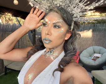 May include: A woman wearing a white, sparkly, halter-style top with a large, silver, snowflake-shaped crown. She has silver glitter makeup on her face and neck.