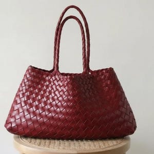 May include: A burgundy tote bag with woven detailing. The bag has two braided handles and sits on a small, round stool. The bag's woven pattern is visible across its body. The background is a plain, off-white wall.